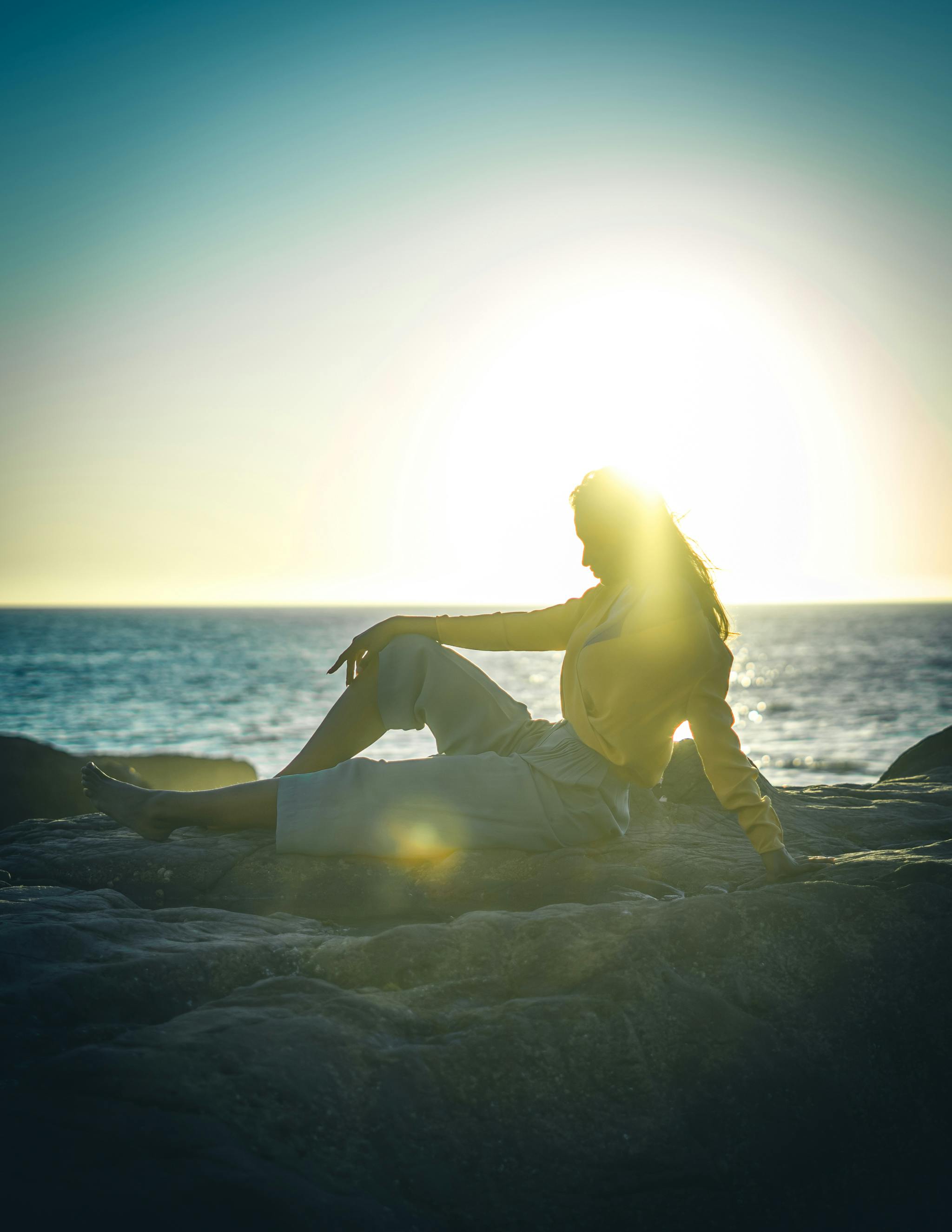 A person sits in a meditation lotus pose on a rock by the sea at sunrise.